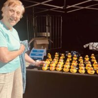 Una mujer mayor posa delante de un stand en el que hay un montón de patos de goma amarillos